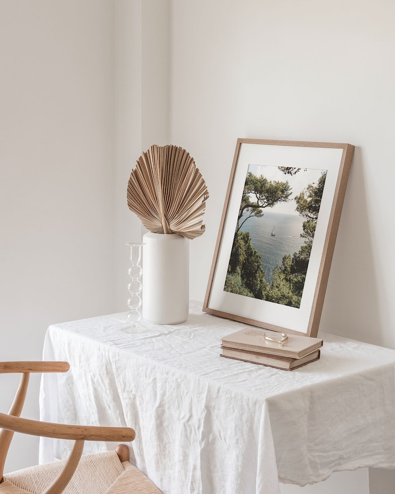K&ouml;nnte beinhalten: Ein wei&szlig;er Tisch mit einer wei&szlig;en Tischdecke aus Leinen, ein Holzstuhl, eine Vase mit einem getrockneten Palmblatt, ein gerahmtes Foto einer Meereslandschaft und zwei &uuml;bereinander gestapelte B&uuml;cher.