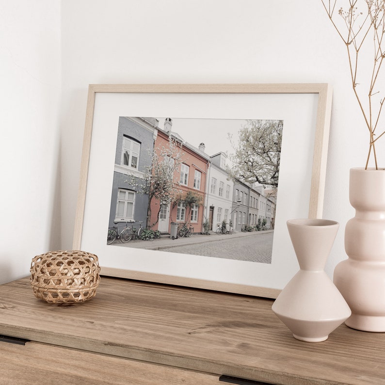 May include: A framed photograph of a street lined with colorful houses in a European city. The houses are painted in shades of gray, red, and white, and have windows and doors. There are trees and bicycles in the street.