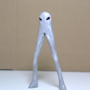 Puede incluir: Figura de alienígena gris con cuerpo delgado y piernas largas. El alienígena tiene una cabeza pequeña con ojos oscuros y una superficie ligeramente texturizada. Las piernas están posicionadas en una postura amplia, dando a la figura una apariencia única.
