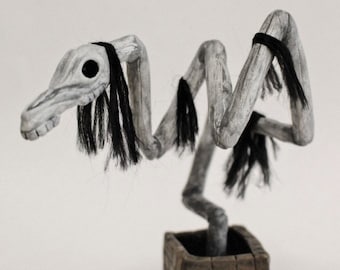 May include: A white, skeletal, and twisted figure with black hair stands on a brown brick base. The figure is made of wood and has a long, thin neck and a small head. It is a unique and eerie sculpture.