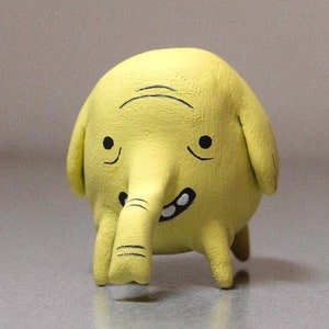 May include: A yellow hand-painted cartoon character with a long nose and a toothy grin. The character is made of clay and has a round body and short legs.