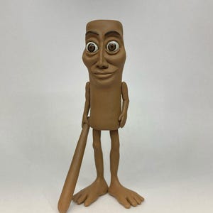 May include: A brown clay figurine of a person with large eyes and a baseball bat. The figurine has a simple, cartoon-like design.