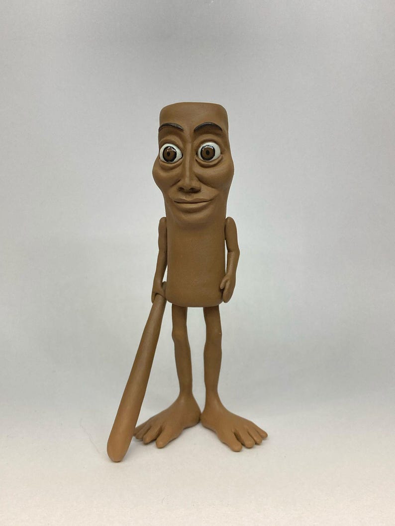 May include: A brown clay figurine of a person with large eyes and a baseball bat. The figurine has a simple, cartoon-like design.