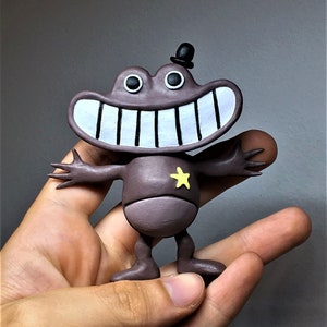 May include: A whimsical, hand-painted figurine of a smiling frog with a top hat and a yellow star on its chest. The frog is a muted purple color with a large white-toothed grin and outstretched arms.