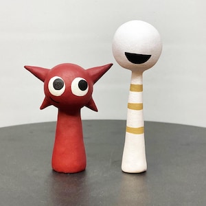 Op de afbeelding: Twee grillige, handgemaakte keramische figuren. Eén is rood met puntige oren en grote ogen. De andere is wit met een rond hoofd, een zwarte glimlach en gouden strepen op de steel. De figuren staan op een donkergrijze ondergrond.