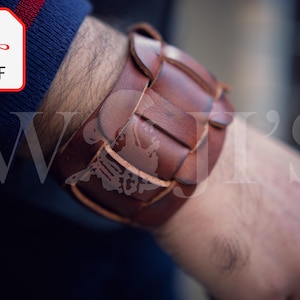 May include: A brown leather bracelet with a woven design. The bracelet is worn on a person's wrist.