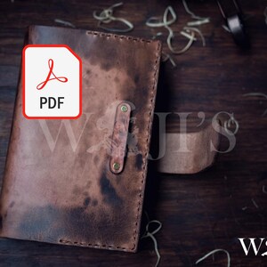 Pode incluir: Um diário de couro castanho artesanal com fecho de correia. O diário apresenta um ícone PDF no canto superior esquerdo. O diário está sobre uma superfície de madeira escura. A marca "W & J's" é visível.