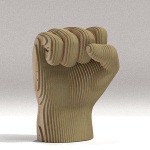 May include: A wooden sculpture of a clenched fist, crafted from layered, light-colored wood. The artwork is a detailed representation of a fist, with visible lines defining each finger and knuckle. The sculpture is set against a neutral background.