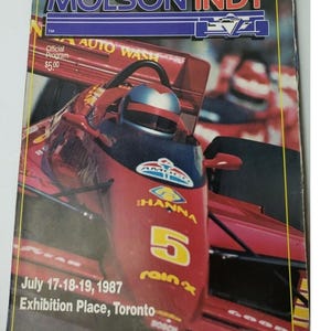 Peut inclure: Un programme de course Molson Indy vintage de 1987, avec une voiture de course rouge portant le numéro 5. Le programme comprend le texte "AUTO WASH" et "HANNA". L'événement a eu lieu à Exhibition Place, Toronto.