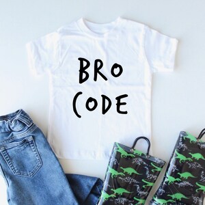 Może przedstawiać: Biała koszulka z napisem "BRO CODE" napisanym czarnym kolorem. Koszulka jest połączona z niebieskimi dżinsami i czarno-zielonymi kaloszami z nadrukiem dinozaurów.