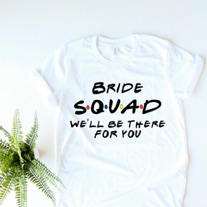 以下が含まれることがあります： 白いTシャツに黒い文字で「Bride Squad」とカラフルなドットと「We'll be there for you」と書かれています。