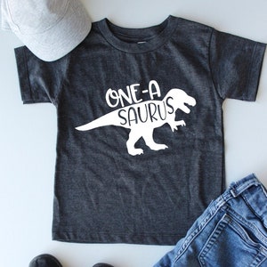 May include: A dark grey short-sleeved t-shirt with a white dinosaur graphic and the text "ONE-A-SAURUS". The shirt is paired with blue jeans, black slip-on shoes, and a grey baseball cap.