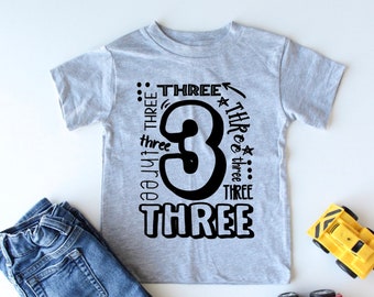 May include: A gray t-shirt with a black number three and the word "three" repeated around it. The shirt is on a white surface with a pair of blue jeans and two toy vehicles.