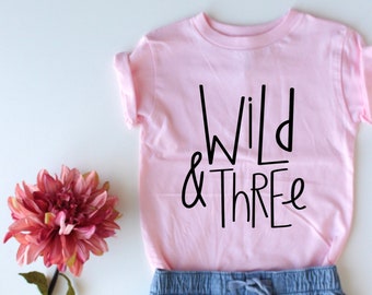 以下が含まれることがあります： ピンクのTシャツに「Wild & Three」という黒い文字が書かれています。Tシャツは青いデニムのショートパンツと組み合わされています。