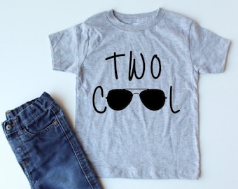 May include: A gray short-sleeved t-shirt with the text "TWO COOL" and a pair of sunglasses printed on it. The shirt is paired with blue jeans.
