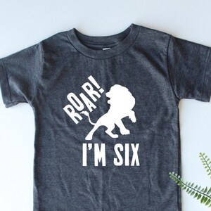 Pode incluir: Uma camiseta cinza escura com uma silhueta de leão branca e o texto "Roar! I'm Six".