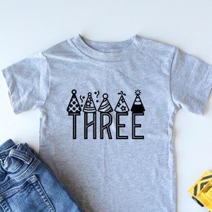 May include: A grey short-sleeved t-shirt with a black graphic of party hats and the word "THREE".