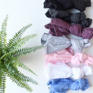 May include: A stack of eight folded t-shirts in various colors, including black, gray, maroon, light gray, pink, white, light blue, and dark blue. The shirts are arranged in a vertical stack, with the black shirt at the top and the dark blue shirt at the bottom. The shirts are all knotted at the bottom.