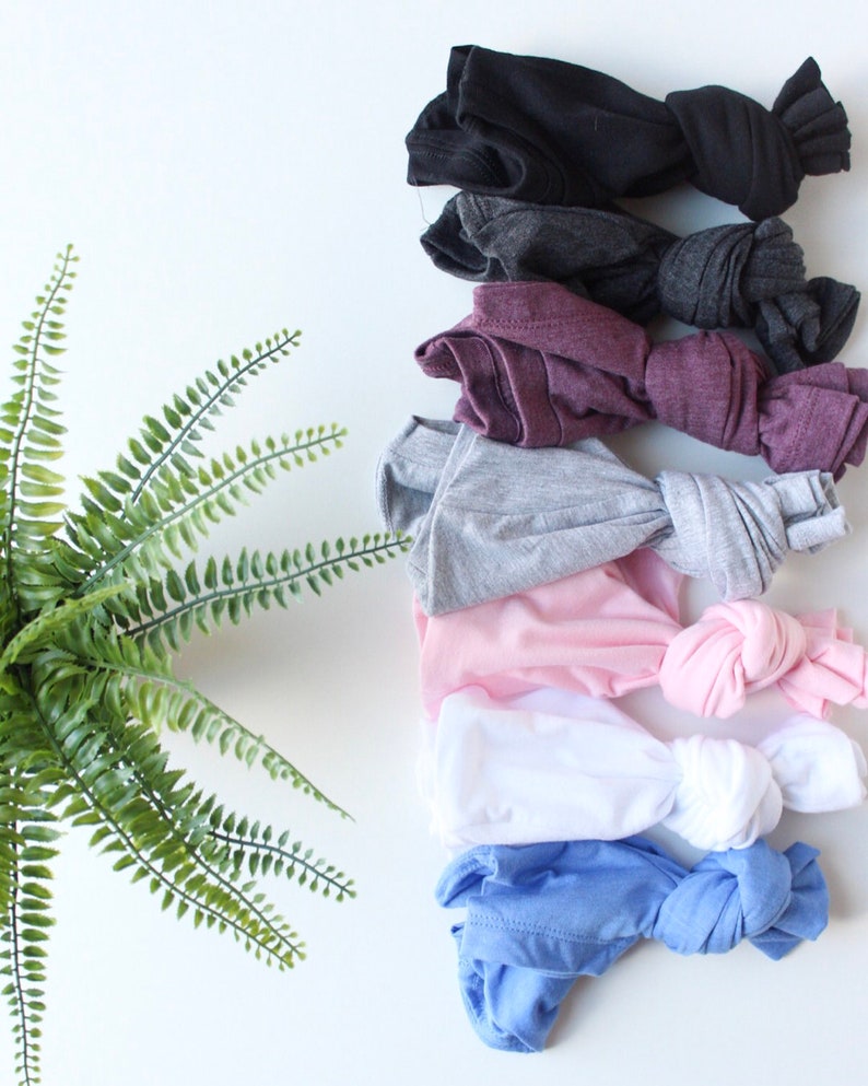 May include: A stack of eight folded t-shirts in various colors, including black, gray, maroon, light gray, pink, white, light blue, and dark blue. The shirts are arranged in a vertical stack, with the black shirt at the top and the dark blue shirt at the bottom. The shirts are all knotted at the bottom.