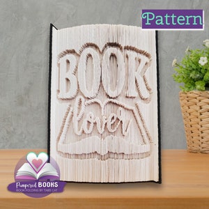 May include: A white book with the words "BOOK LOVER" folded into the pages. The book is standing upright on a wooden surface. The book is a craft project.