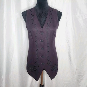 May include: A dark purple vest with a V-neck and black decorative trim. The vest has a button-up front and a tailored fit. The trim is a wavy, embroidered design. The vest is displayed on a mannequin.