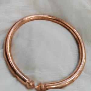 May include: A copper-coloured, open bangle bracelet. The bracelet is a thick, rounded band with a slight gap at the front. The ends of the bracelet have a geometric design. The bracelet is displayed on a white, textured surface.