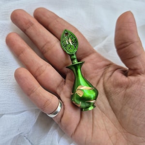 May include: A small, green perfume bottle with a leaf-shaped top and gold accents. The bottle has a rounded shape and is held in a hand. The background is a white, textured fabric. The bottle is approximately 7.6 cm tall.
