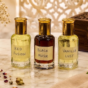 May include: Three glass perfume bottles with gold caps, each featuring a rectangular label. The left bottle contains light yellow liquid labelled Red Fusion, the centre bottle has dark brown liquid labelled Black Musk, and the right bottle contains light yellow liquid labelled Vanilla Lust.