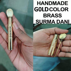 May include: A gold-colored brass surma dani, a traditional Indian eye cosmetic applicator, with a round top and a textured body. The surma dani is held in a hand.