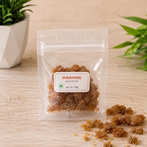 May include: A clear plastic resealable pouch containing brown, crystalline pieces of asafoetida. The white label on the front reads HIRAHING (ASAFOETIDA) with a green square symbol and Net Wt. 10g. Several loose pieces are scattered nearby.
