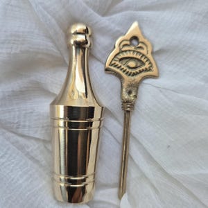 May include: A gold metal bottle stopper with a decorative eye design. The stopper has a long, thin metal rod that fits inside the bottle.