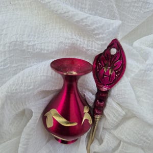 May include: A small, crimson red vase with gold accents and a matching gold-coloured, ornate, decorative stick. The vase and stick are on a white fabric background.