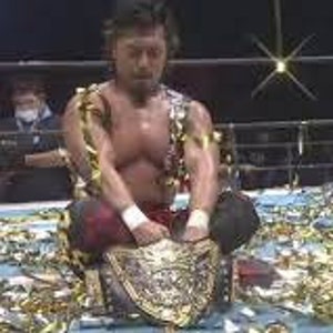 Puede incluir: Un luchador con un traje de lucha rojo y negro se sienta en un escenario cubierto de confeti dorado. Está sosteniendo un cinturón de campeonato con un diseño dorado y negro. El cinturón tiene una hebilla grande y adornada.