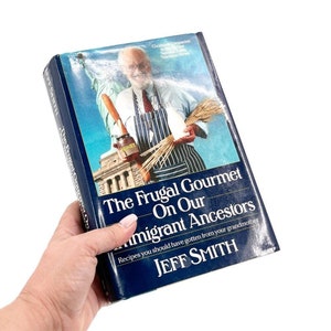 Puede incluir: Un libro de cocina de tapa dura azul titulado "The Frugal Gourmet On Our Immigrant Ancestors" de Jeff Smith. La portada presenta a un hombre con un delantal a rayas sosteniendo una hogaza de pan y un tarro de miel.