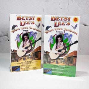 May include: Two VHS tapes of "Betsy Lee's Ghost Town Jamboree", one labeled "Volume 1" and the other "Volume 2". Both tapes feature a woman playing a guitar in front of a Western town backdrop. The tapes are in their original packaging.