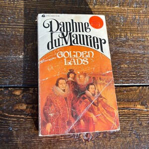Pode incluir: Um livro de bolso vintage intitulado "Golden Lads" de Daphne du Maurier. A capa laranja apresenta um retrato de época. O nome do autor está em letras pretas grandes. O livro mostra sinais de uso.