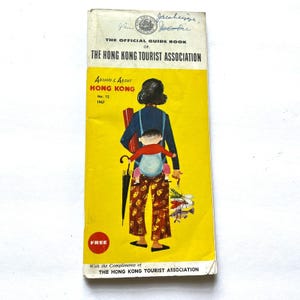 Puede incluir: Guía turística vintage de Hong Kong de 1967. La portada amarilla presenta una ilustración de una persona que lleva a un niño, sosteniendo un paraguas y una bolsa de artículos. El título de la guía es "Around & About Hong Kong."