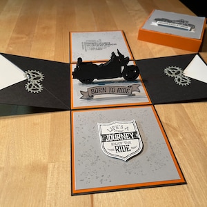 Könnte beinhalten: Eine schwarz-weiße Pop-up-Karte mit einem Motorrad-Design. Die Karte ist geöffnet und zeigt ein schwarzes Motorrad mit den Worten "Born to Ride" auf einem Banner. Die Karte hat auch einen grauen Hintergrund mit den Worten "Life's a Journey, Enjoy the Ride" auf einem Schild.
