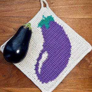 Könnte beinhalten: Ein handgefertigter, quadratischer, cremefarbener Topflappen aus Häkelarbeit mit einem lila Aubergine-Design und einem grünen Stiel. Eine frische, dunkelviolette Aubergine liegt neben dem Topflappen. Der Topflappen hat eine Schlaufe zum Aufhängen.
