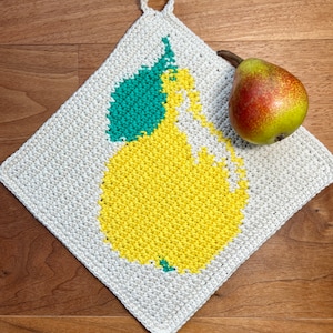 Könnte beinhalten: Ein handgefertigter, cremefarbener Topflappen aus Häkelarbeit mit einem gelben Birnenmuster und grünen Blättern. Eine echte Birne mit rot-grüner Schale liegt auf dem Topflappen. Der Topflappen hat eine Schlaufe zum Aufhängen und liegt auf Holz.