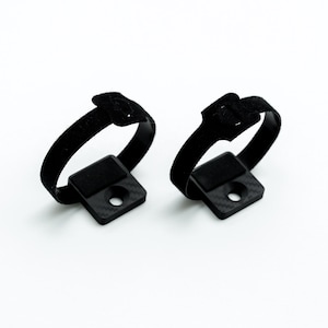 May include: Two black velcro straps with square, textured bases. Each base has a central hole. The straps are designed to secure items. The background is plain white.