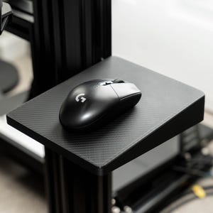 Puede incluir: Un ratón de ordenador negro con textura de fibra de carbono se encuentra sobre una alfombrilla de ratón negra. La alfombrilla de ratón está unida a un soporte de metal negro.