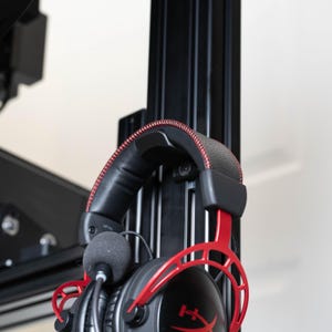May include: Red and black gaming headset with a microphone hanging from the side. The headset is hanging on a black metal stand.
