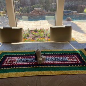 May include: A colorful table runner with a geometric pattern in shades of red, blue, green, yellow, and black. The runner is laid out on a wooden table with two white chairs and a white ceramic jar with two white candles.