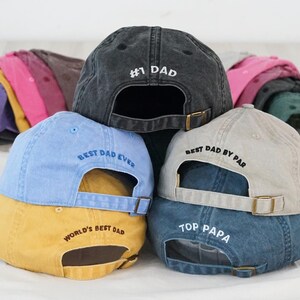 May include: Several baseball caps in shades of blue, yellow, and grey, stacked together. Each cap features embroidered text, including "#1 DAD", "BEST DAD EVER", and "TOP PAPA". Adjustable straps are visible.