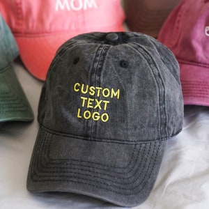 Può includere: Cinque cappellini da baseball di diversi colori con testo ricamato. I cappellini sono verde, rosa, marrone, bordeaux e nero. Il testo sui cappellini recita "DAD EST. 2024", "BEST BONES MOM", "CUSTOM TEXT LOGO" e una silhouette di un cane.