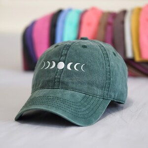 Op de afbeelding: Een donkergroene baseballpet met een wit geborduurd ontwerp van een wassende maan en volle maanfasen.