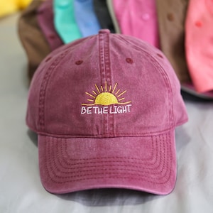 Könnte beinhalten: Eine marineblaue Baseballkappe mit einer aufgestickten gelben Sonne und dem Text "BE THE LIGHT" in Weiß.