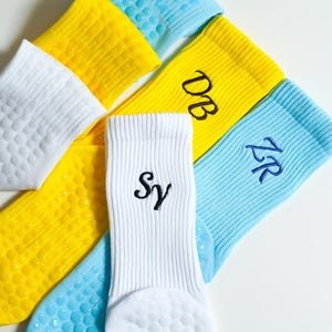 Pode incluir: Uma coleção de meias caneladas em amarelo, branco e azul claro. As meias apresentam iniciais bordadas, incluindo DB, SY e ZR. As solas das meias são cobertas com pequenos pontos circulares de silicone para maior aderência.