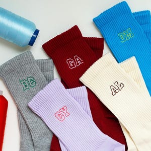 Puede incluir: Calcetines acanalados surtidos en varios colores, incluyendo gris, rojo, azul y crema. Cada calcetín presenta iniciales bordadas en colores contrastantes. Se ven carretes de hilo azul y naranja, lo que sugiere un tema artesanal.